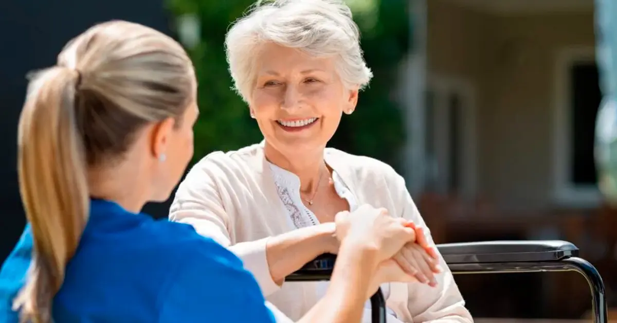 Smiling senior patient sitting on wheelchair with nurse supporting her. Doctor looking at elderly patient on a wheelchair in the garden. Nurse holding hand of mature woman outside care home.